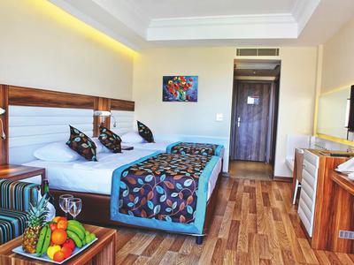 Helles Hotelzimmer mit Doppelbett, bunten Kissen, Holzfußboden und Früchtekorb auf Tisch neben Sessel.