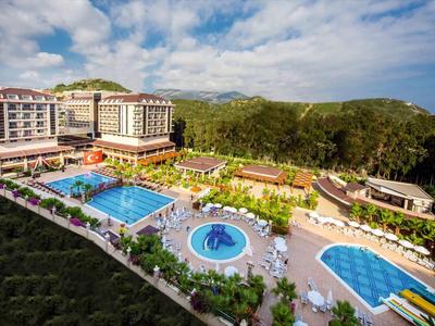 Große Hotelanlage mit mehreren blau gefüllten Schwimmbecken, umgeben von grünen Hügeln und bewölktem Himmel.