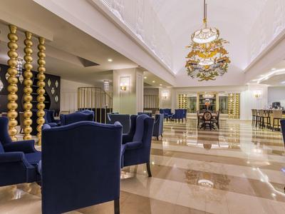 Una luminosa hall d'hotel con poltrone blu scuro, pavimento in marmo beige e colonne dorate decorative.