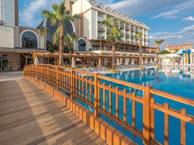 Hotel di lusso con piscina e passerella in legno sotto un cielo limpido e palme.