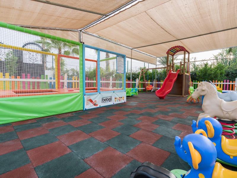 Zadaszony plac zabaw dla dzieci z zjeżdżalnią i konikami na biegunach obok siatki do siatkówki.