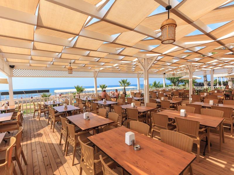 Duży zadaszony taras z drewnianymi stołami i widokiem na morze w słoneczny dzień.