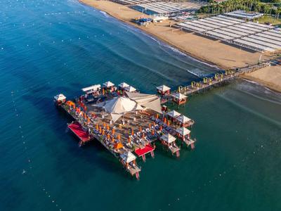 Vista aérea de un muelle largo con edificios y barcos, rodeado de mar azul y playa.