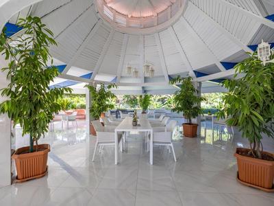 Área exterior cubierta con mesas y sillas blancas, rodeada de grandes plantas en macetas y vista al mar.
