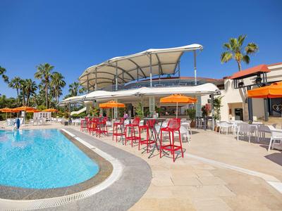 Área de piscina del hotel con sombrillas, sillas rojas y palmeras bajo cielo despejado.