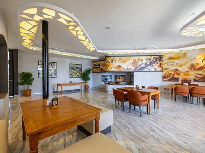 Área de salón luminosa y moderna en hotel con mesas de madera, sillones y luces decorativas en el techo.