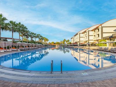 Gran piscina redonda con agua azul clara, rodeada de palmeras y edificios de hotel bajo cielo azul.