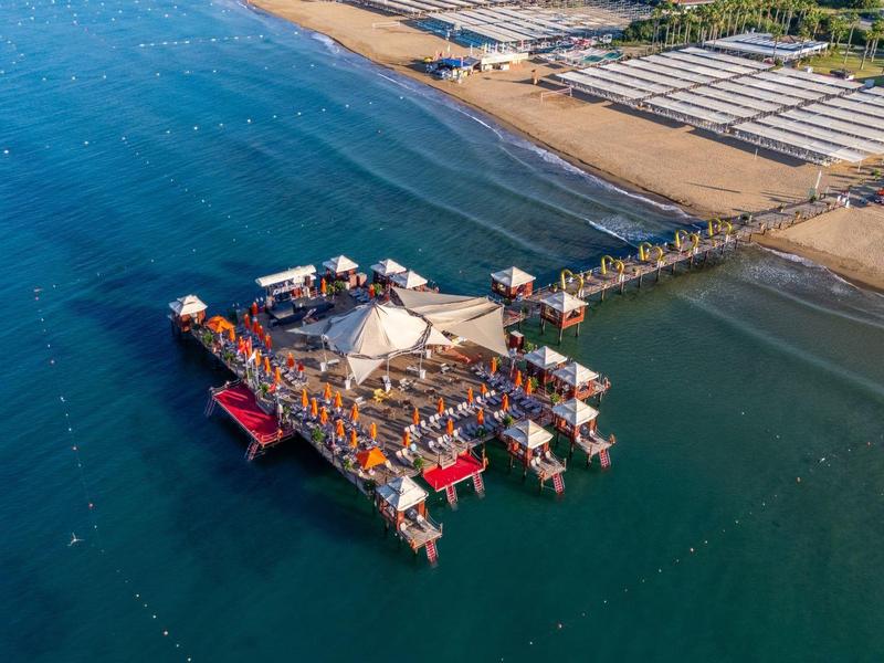 Vista aérea de un muelle largo con edificios y barcos, rodeado de mar azul y playa.