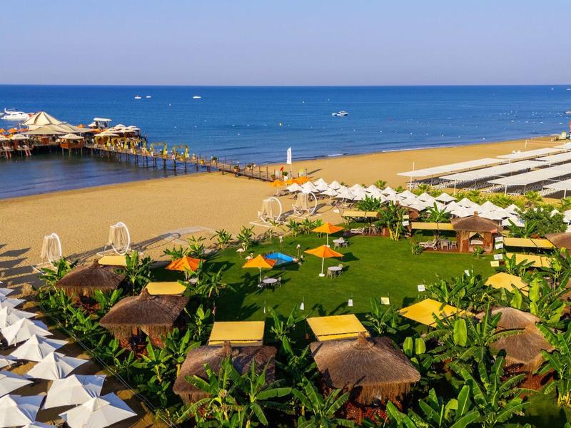 Vista de una playa con sombrillas, cabañas y un muelle de madera en el mar con cielo despejado.