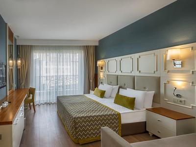 Modern hotel room with double bed, wood furniture, teal walls, and sheer curtains lighting the space.