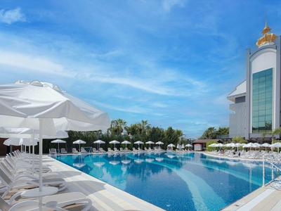 Großer Pool mit blauer Wasseroberfläche, weißen Liegestühlen und Sonnenschirmen vor modernem Hotelgebäude.