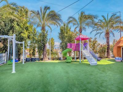 Outdoor playground with slide, swings, and palm trees on a green artificial turf.
