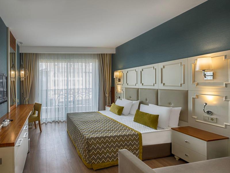 Modern hotel room with double bed, wood furniture, teal walls, and sheer curtains lighting the space.