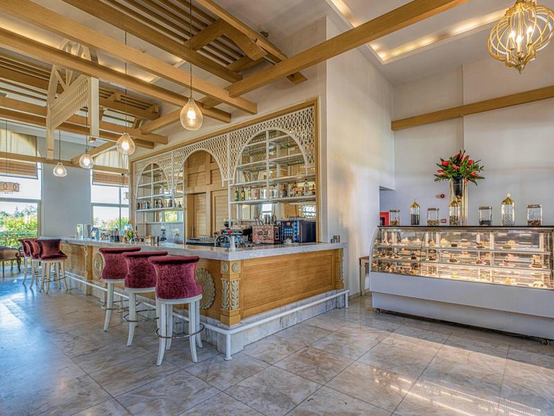 Bright hotel lobby with wooden ceiling, bar counter, velvet stools, and display case of pastries.