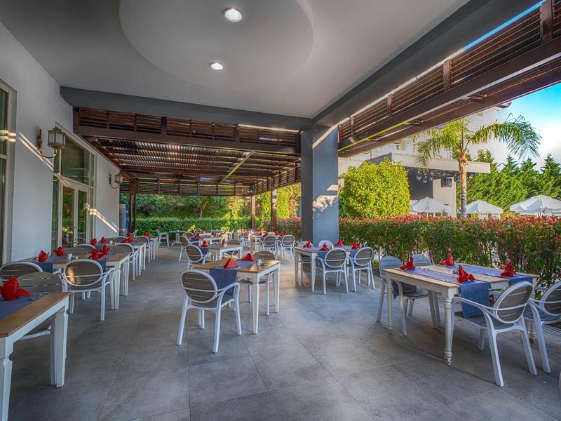 Outdoor hotel dining area with tables, chairs, and greenery under wooden pergola.