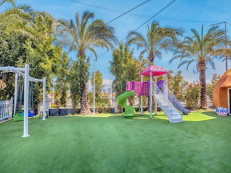 Outdoor playground with slide, swings, and palm trees on a green artificial turf.