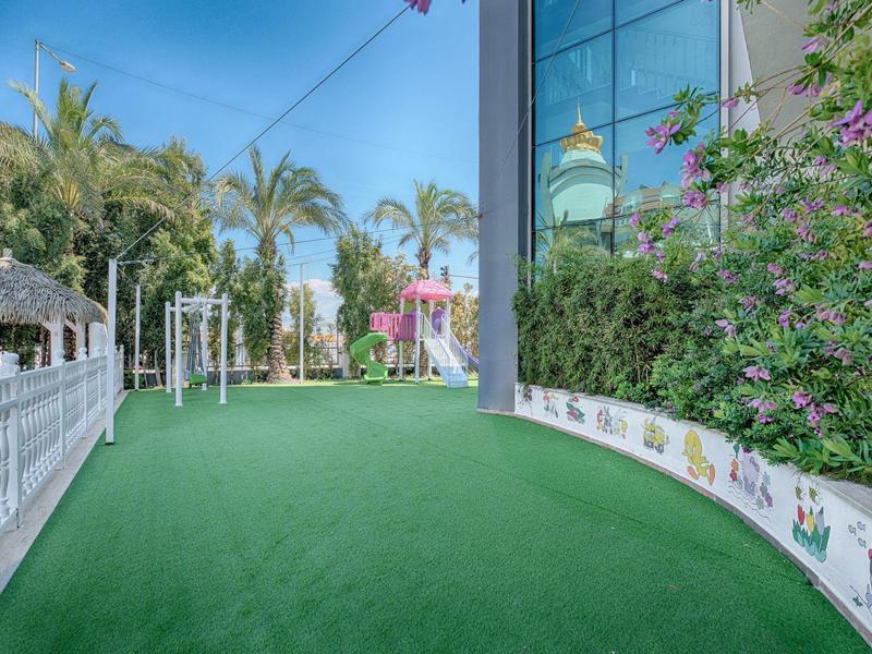 Outdoor space with green artificial turf, decorative flowerbed, palm trees, and a white gazebo under clear sky.