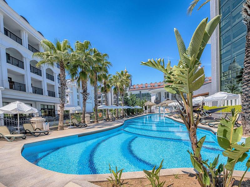 Outdoor swimming pool surrounded by lounge chairs and palm trees next to a hotel under a clear blue sky.