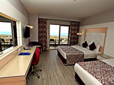 Světý hotelový pokoj se dvěma postelemi, stolem a balkónem s výhledem na moře.