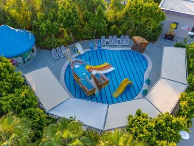 Kruhový venkovní bazén s vodní skluzavkou, nafukovacími hračkami a lehátky obklopený stromy.