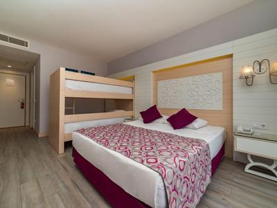 Hotelový pokoj s manželskou postelí, palandou, dřevěnou podlahou a moderním nástěnným osvětlením.