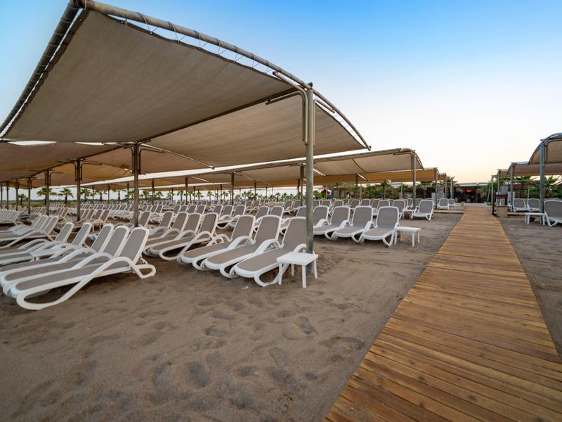 Pláž s lehátky pod zastíněnými přístřešky a dřevěnou chodníkem při soumraku.