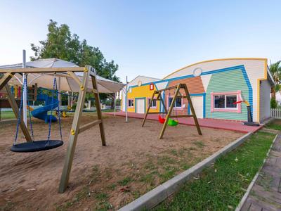 Speeltuin met schommels voor kleurrijke, gelijkvloerse vakantiehuisjes bij heldere lucht.