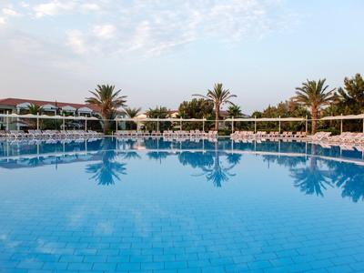 Helderblauw zwembad met palmbomen en ligstoelen bij een tropisch resort onder een gedeeltelijk bewolkte lucht.
