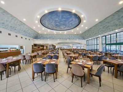 Grote, lichte eetzaal met veel tafels en moderne stoelen, ronde plafondlamp en grote ramen.