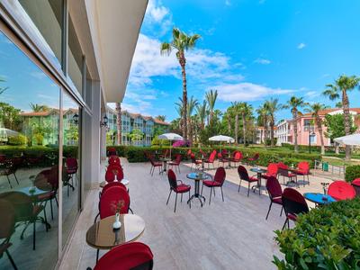 Terras met rode stoelen en tafels voor een hotel met palmbomen en blauwe lucht.