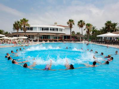 Groot hotelzwembad met veel mensen die in een cirkel spetteren, omgeven door palmbomen en parasols.