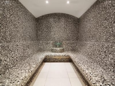 Moderne stoombadkamer met mozaïektegels op muren en banken, heldere plafondlampen en betegelde vloer.