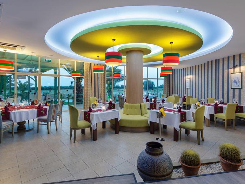 Moderne restaurantinterieur met ronde plafondlamp, gele stoelen en uitzicht op buiten.