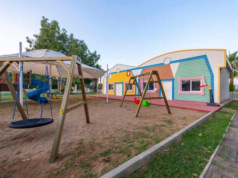 Kleurrijke speeltuin naast een vakantiehuis met schommels en zandbak onder een zonnige lucht.