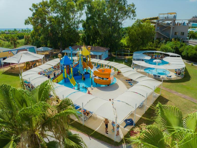 Buitenspeelplaats met waterglijbanen en watersproeiers omringd door schaduwrijke zithoeken.