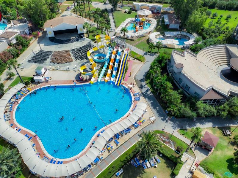 Luchtfoto van een waterpark met groot zwembad, glijbanen en ronde podiumruimte omgeven door groen.