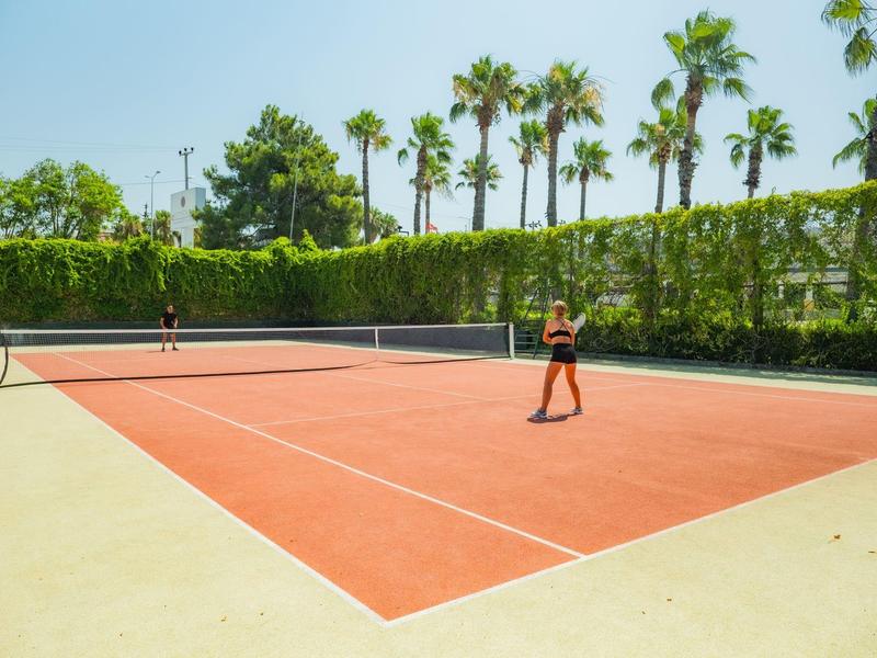 Twee mensen spelen tennis op een buiten rode kleibanen omgeven door palmbomen en groen.
