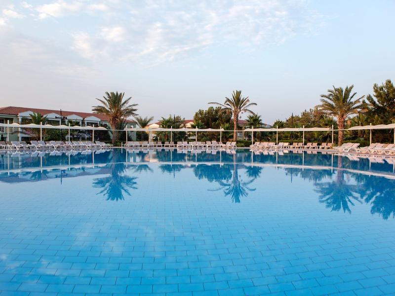 Helderblauw zwembad met palmbomen en ligstoelen bij een tropisch resort onder een gedeeltelijk bewolkte lucht.