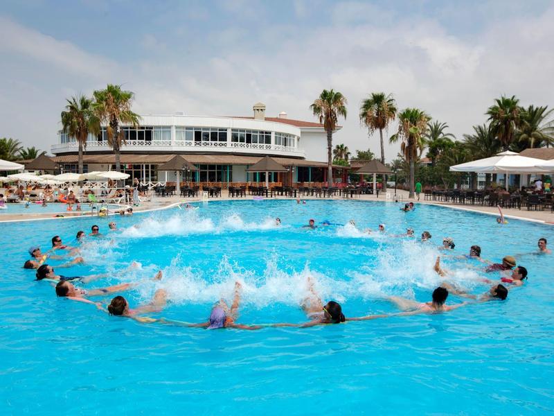 Gasten genieten van een zonnige dag in een groot rond hotelzwembad met palmbomen en een wit gebouw.