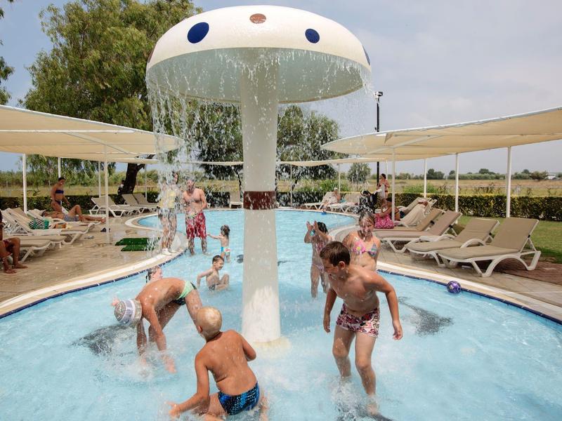Kinderen spelen in een ondiep zwembad onder een paddenstoelvormige waterpartij bij een zonnig resort.