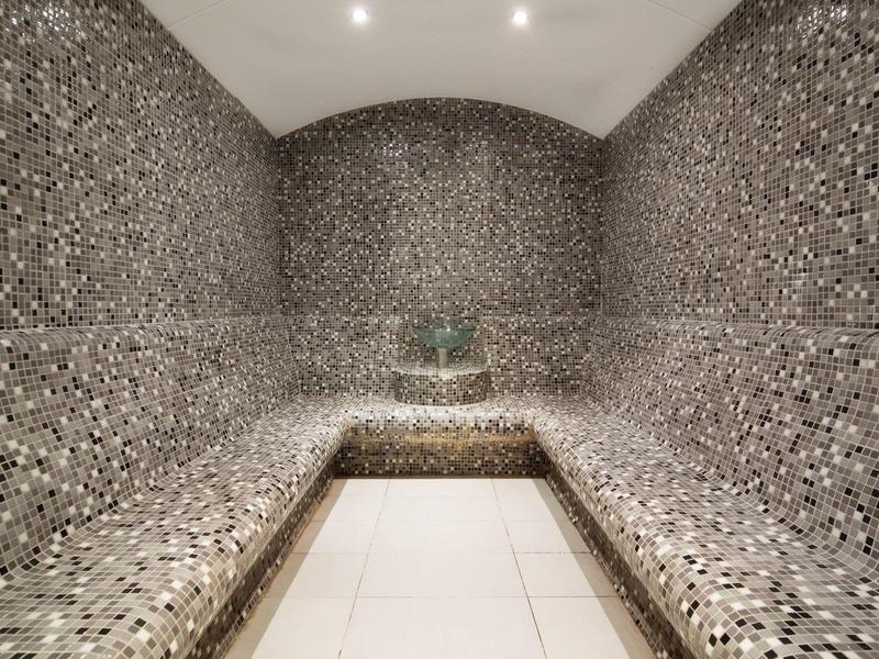 Moderne stoombadkamer met mozaïektegels op muren en banken, heldere plafondlampen en betegelde vloer.