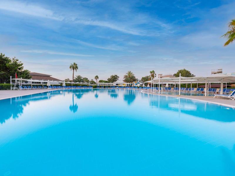 Groot buitenzwembad met helderblauw water, omgeven door ligstoelen en palmbomen.
