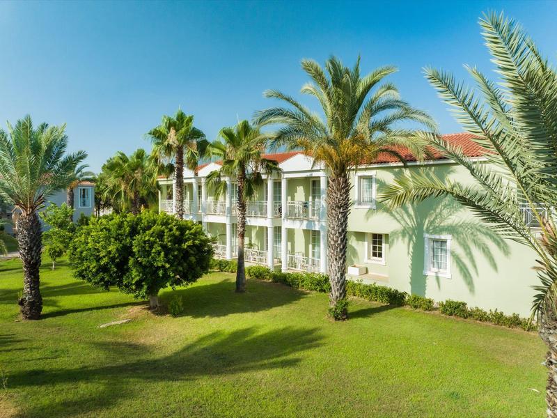 Hotelgebouw met balkons omgeven door palmbomen en groen gras onder een heldere blauwe lucht.
