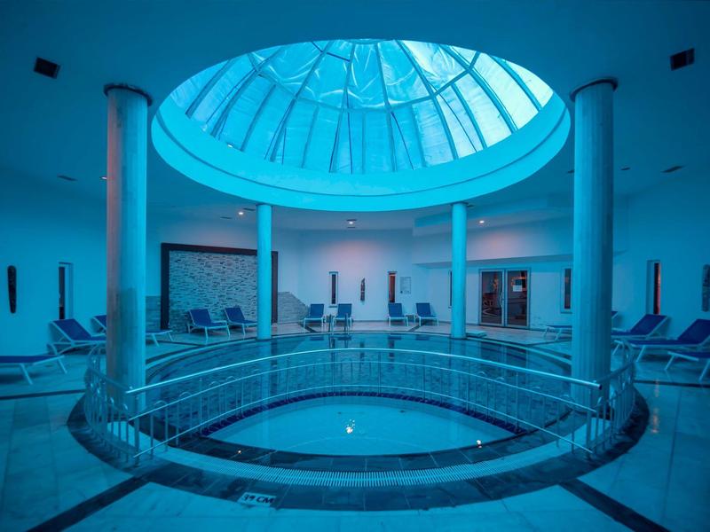 Overdekt spa zwembad met grote glazen koepel en ligstoelen eromheen in een rustgevende omgeving.