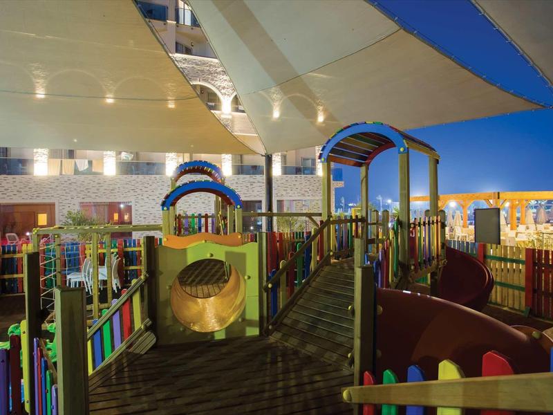 Aire de jeux colorée et sécurisée dans un hôtel, éclairée le soir sous une voile tendue.