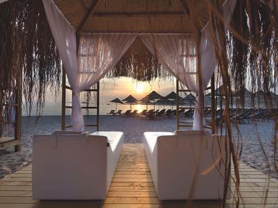 Two white loungers under a thatched roof overlooking the beach and sun umbrellas at sunset.