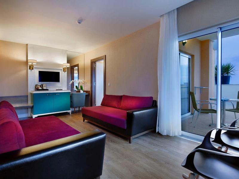 Modern hotel room lounge with red sofas, TV, and access to balcony with table and chairs.
