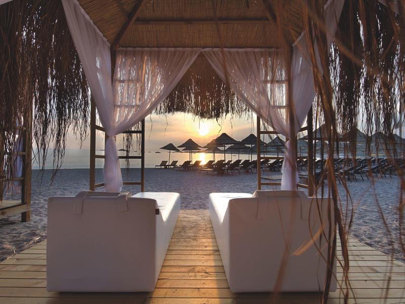 Two white loungers under a wooden canopy on the beach with a view of sunset and umbrellas.