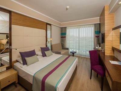 Modernes Hotelzimmer mit Doppelbett, violetten Akzenten, Schreibtisch, Sessel und großen Fenstern mit Vorhängen.