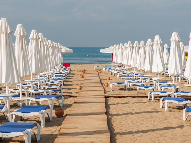 Strand mit symmetrisch angeordneten Liegestühlen, geschlossenen weißen Sonnenschirmen und Blick aufs Meer.